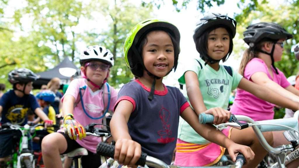 Children with helmets riding bicycles outdoors in a tree-surrounded cycling event.