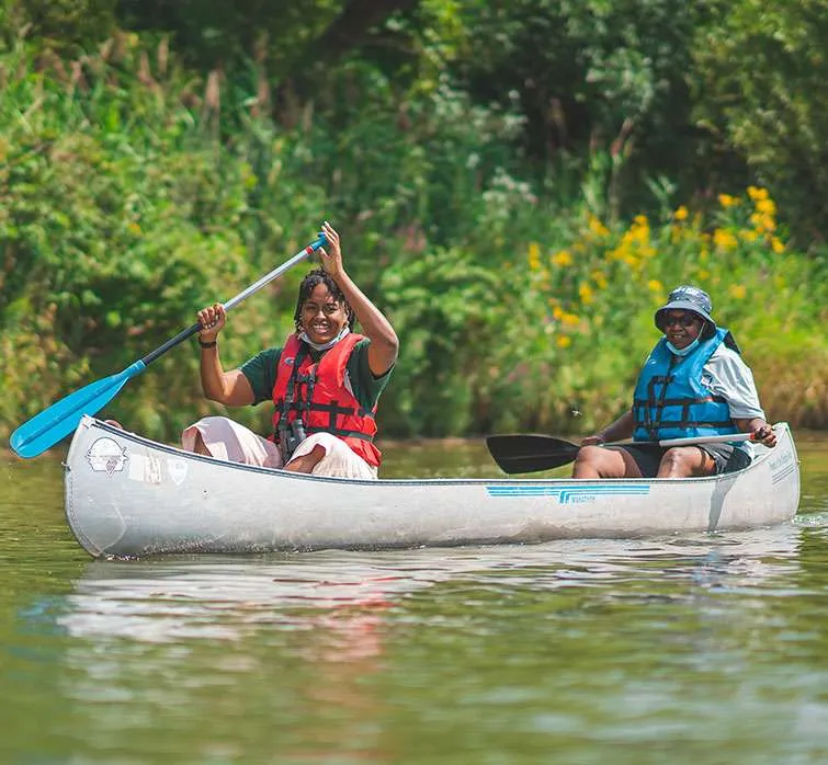 Two people in life vests canoeing on a calm river with green vegetation and yellow flowers nearby.