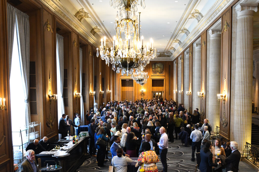 A large group in an ornate ballroom with chandeliers, mingling at a social event near a bar.