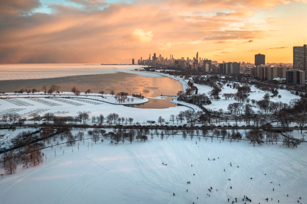 Hermosa fotografía aérea del atardecer de la orilla del lago de Chicago sobre Cricket Hill y el puerto de Montrose con coloridas nubes rosadas y amarillas reflejándose en el agua en una abertura en el hielo que cubre el lago Michigan.