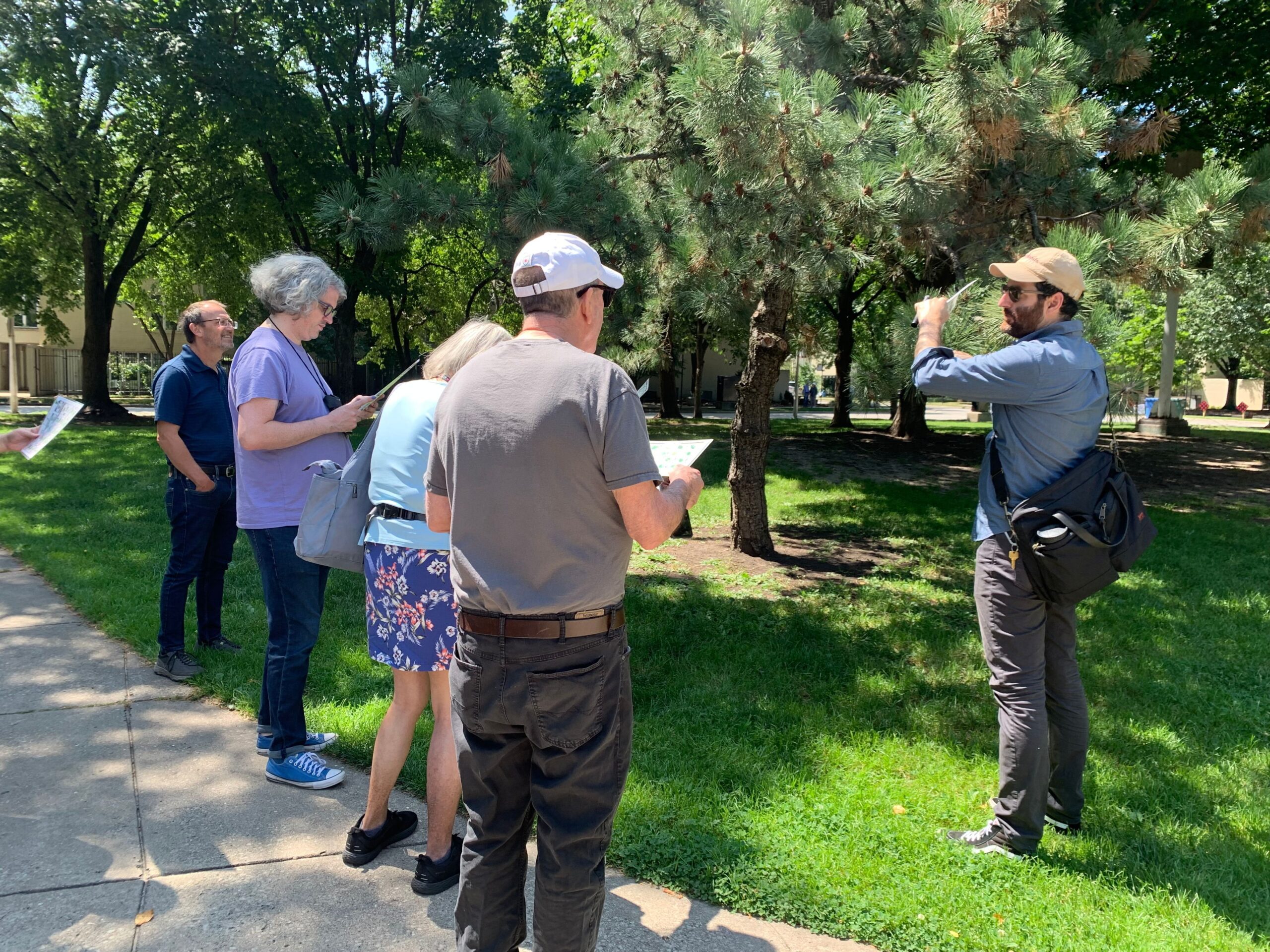 A gentleman standing on the green pasture of a park teaching about a tree to a group of people