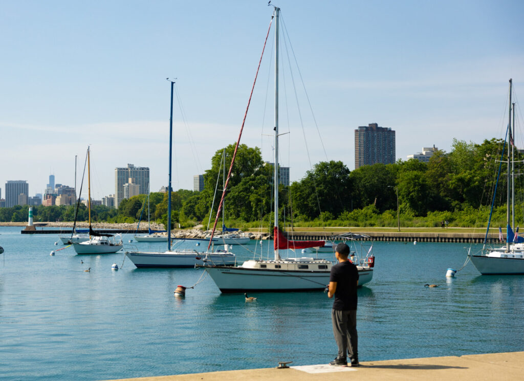 A man fishing on the edge of a marina with boats passing under a clear blue sky.