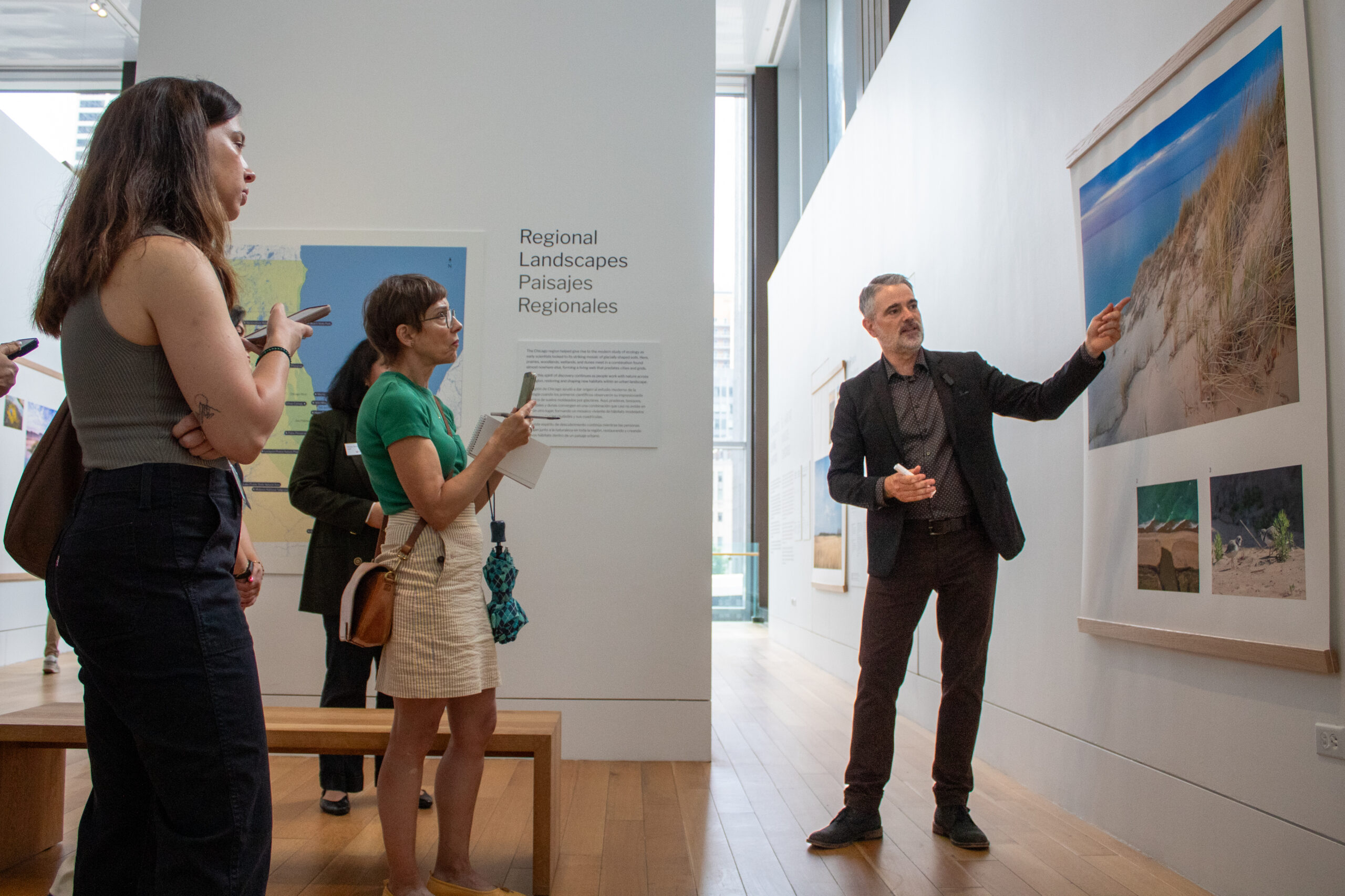 Art gallery visitors observes a presenter pointing to a framed landscape photograph of sand dunes and Lake Michigan.
