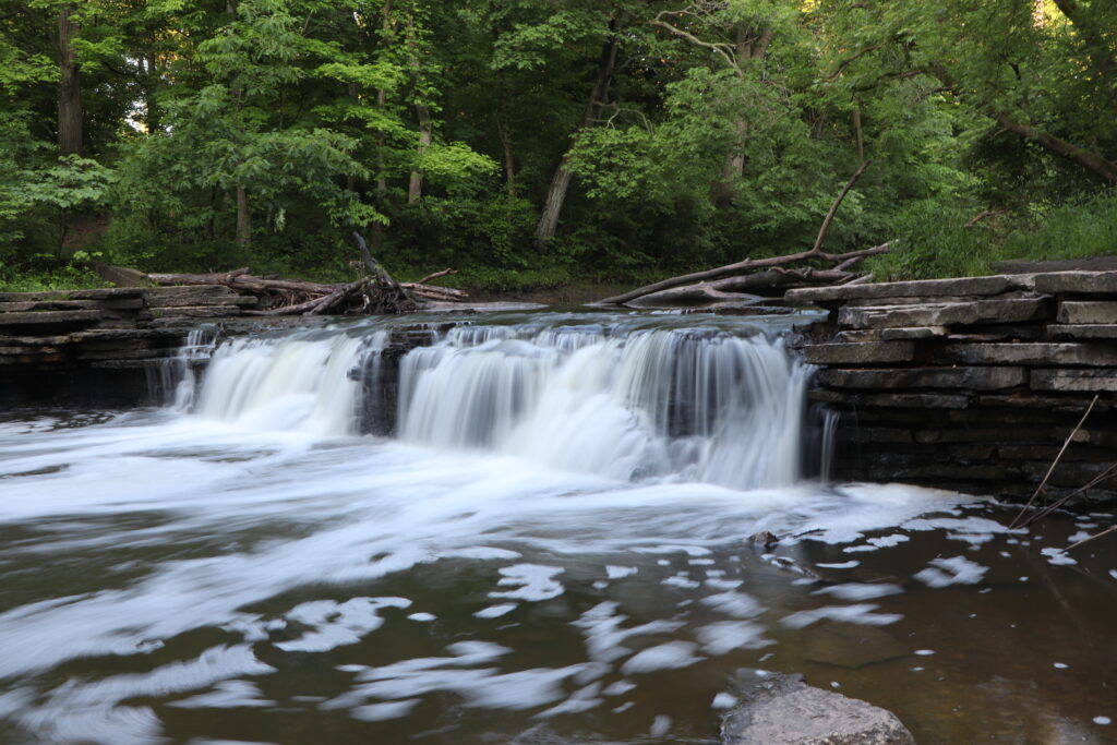 Stream leading to small waterfall over stacked stones with with trees in the background.
