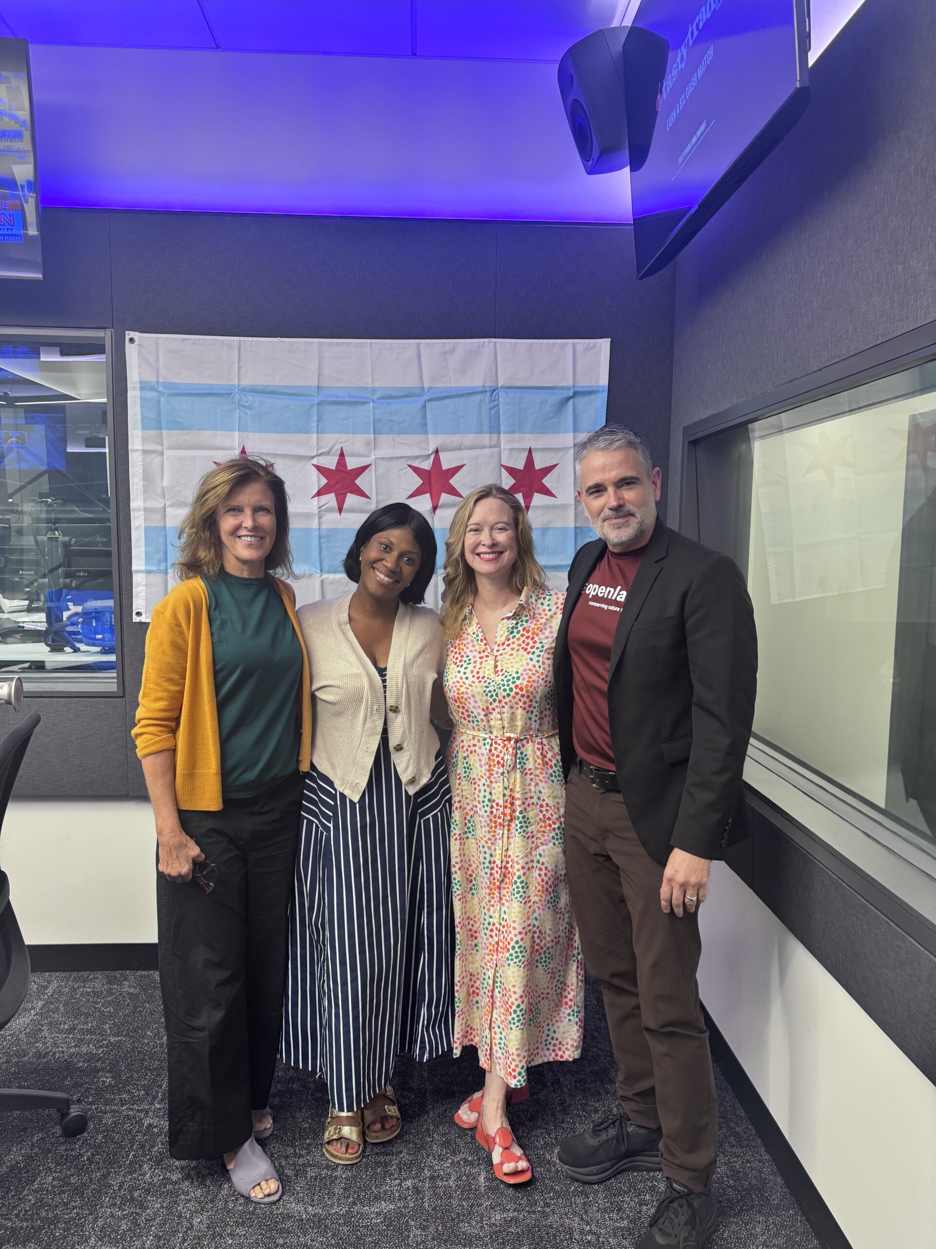 Four adults pose for photo in radio studio, smiling in front of Chicago flag.