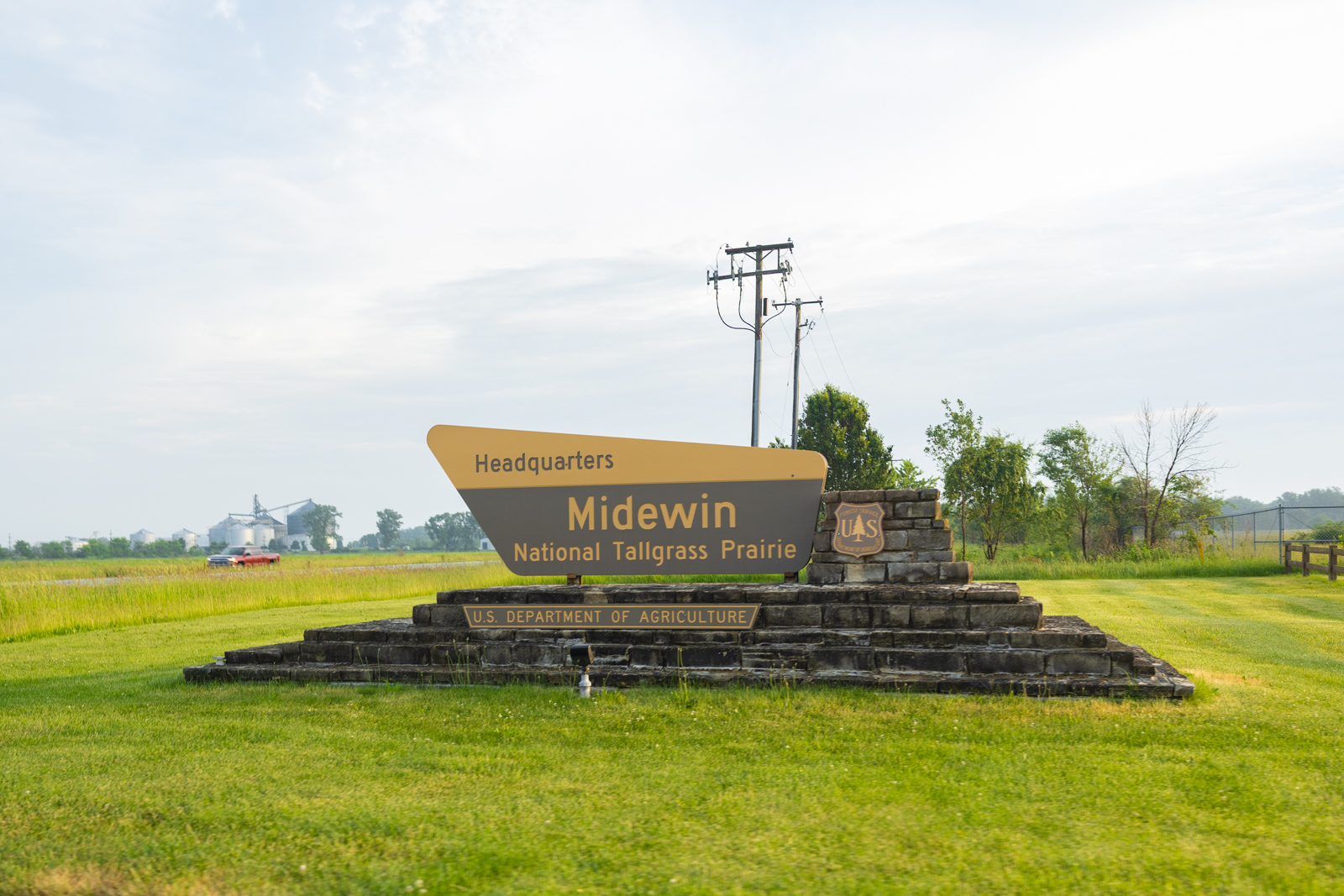 Midewin National Tallgrass Prairie headquarters sign on a stone monument, set in a mowed green lawn with utility poles behind.