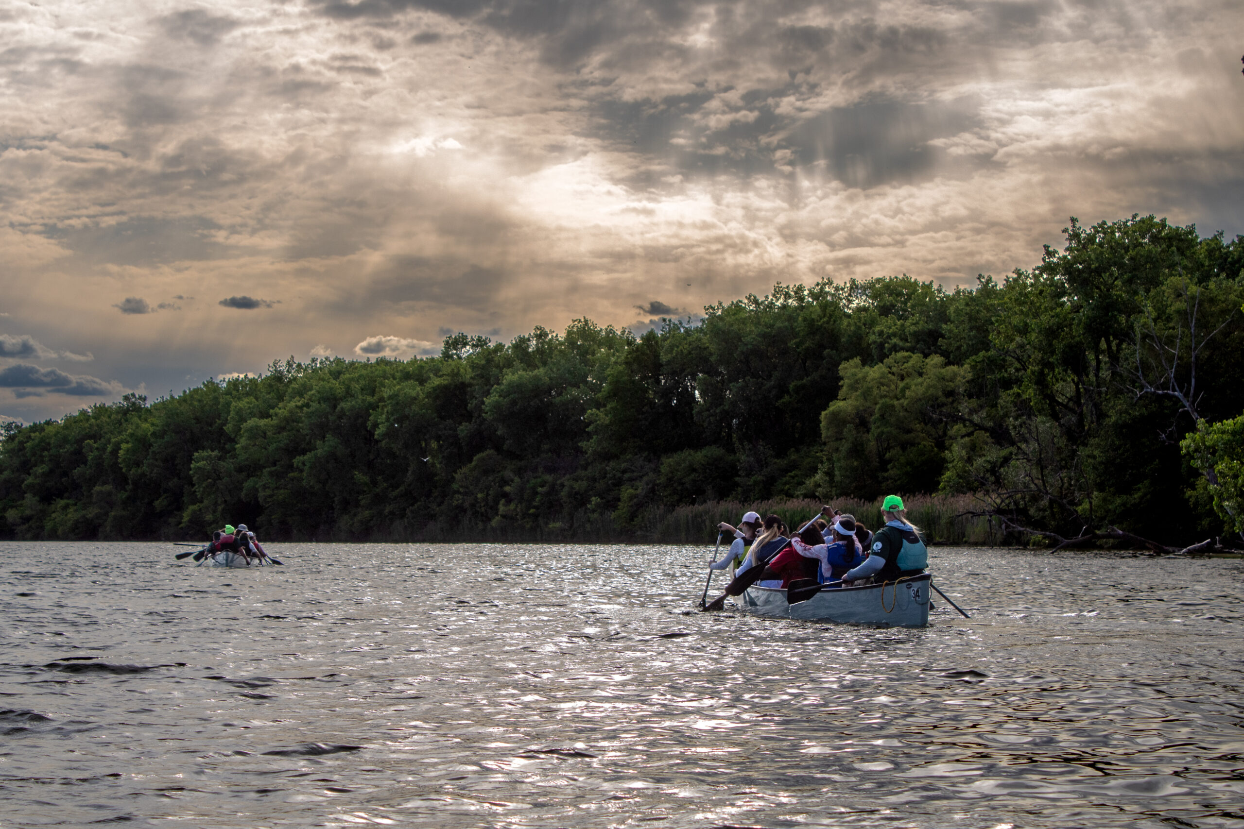 Two canoes paddling on river under cloudy sky with rays of light coming through.
